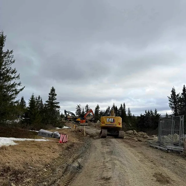 Gravemaskiner fra Maskinkraft AS arbeider med terrengbearbeiding i skogsområde, omgitt av fjell og trær under overskyet himmel.