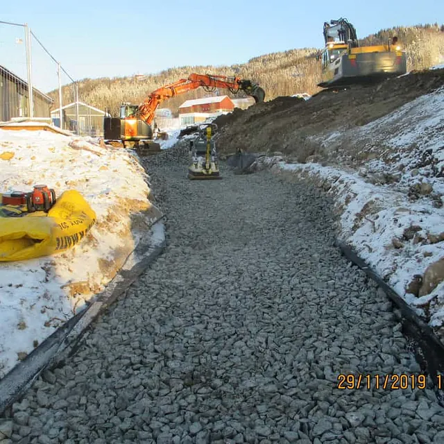 Grøft fylt med pukk under anleggsarbeid i snødekt terreng, gravemaskiner i bakgrunnen.