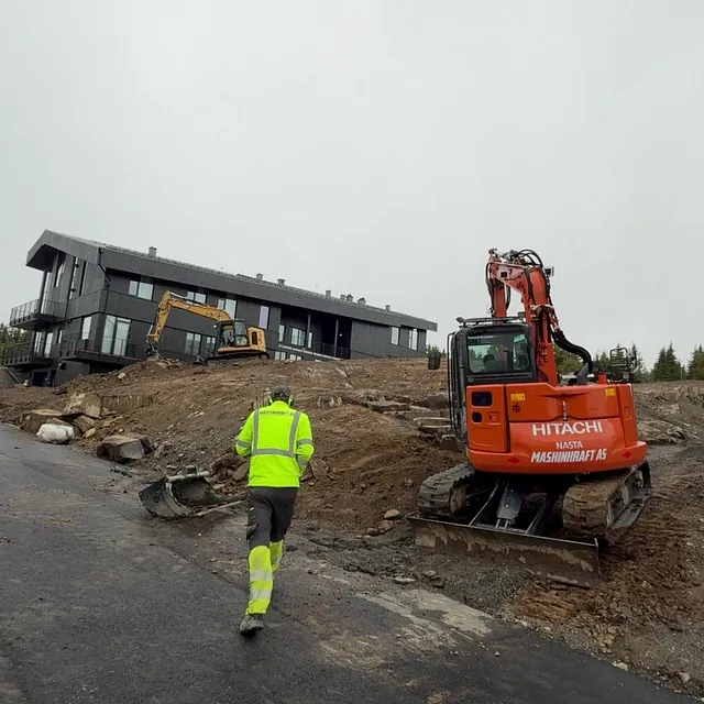 Anleggsarbeider går opp en bratt bakke mot nybygde boliger mens gravemaskiner utfører terrengarbeid.