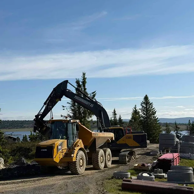 Gravemaskin laster masser i stor dumper på fjellområde med rørdeler og utsikt over vann og fjell.