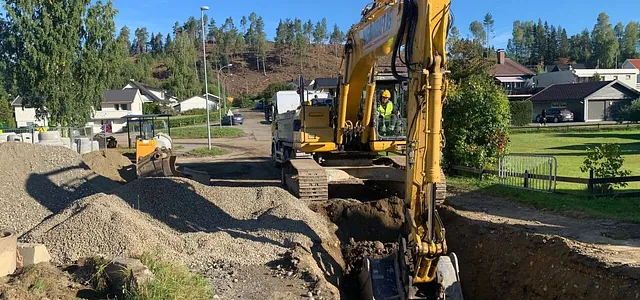 Gravemaskin graver grøft i boligområde på solfylt dag, omgitt av pukkhauger og anleggsmaskiner.
