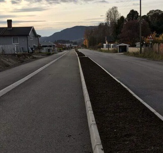 Nyanlagt vei med asfaltert kjørebane, kantstein og midtrabatt fylt med jord, omgitt av boliger og fjell i bakgrunnen.