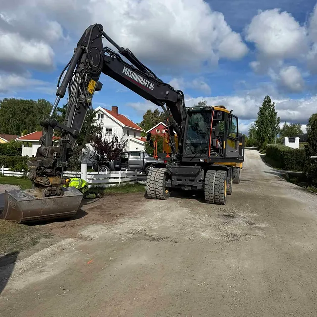 Gravemaskin parkert i boligområde med anleggsarbeider som jobber ved siden av på gruslagt vei.