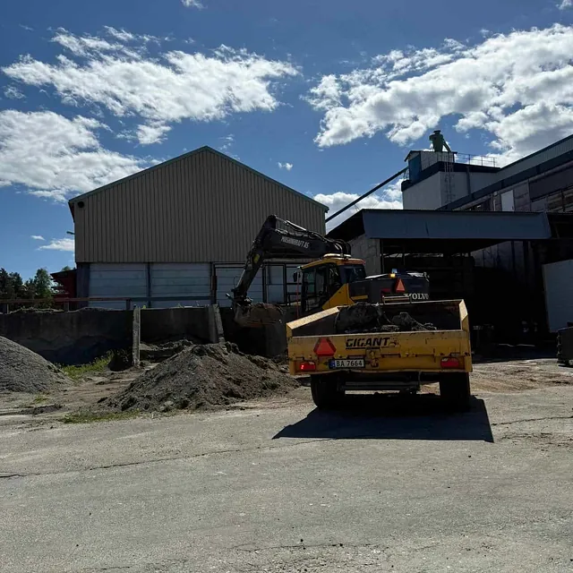 Gravemaskin fra Maskinkraft AS laster masser i henger ved industriområde med bølgeblikksbygning og klar blå himmel.