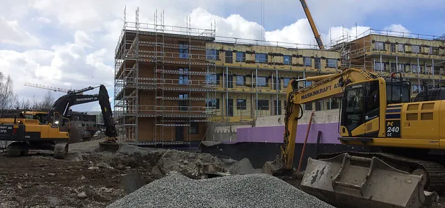 To gravemaskiner arbeider foran et nybygg med stillas, i en boligblokk under oppføring.