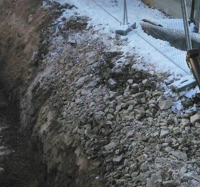 Dyp grøft med stein og gravemaskin, langs nettinggjerde i snødekt boligområde.