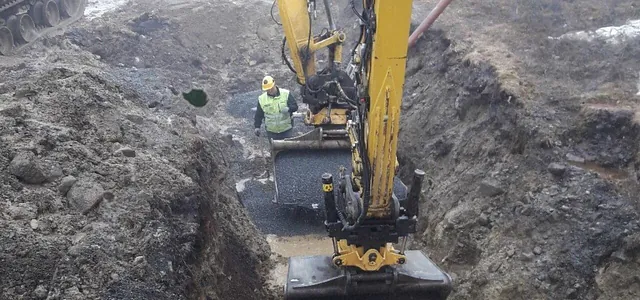 Gravemaskin legger ut pukk i grøft mens en anleggsarbeider følger med nede i grøften.