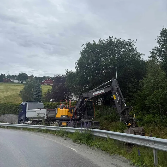 Gravemaskin fra Maskinkraft AS arbeider langs veikant med lastebil parkert ved siden av på svingete asfaltvei.