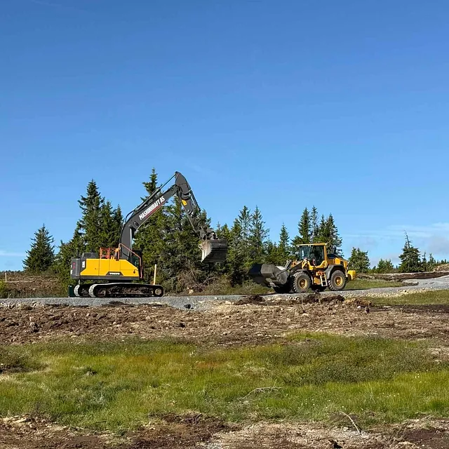 Gravemaskin og hjullaster jobber sammen med massetransport i skogslandskap på steinlagt underlag.