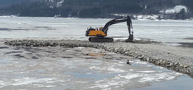 Gravemaskin arbeider på steinfylling utover islagt vann, med snødekte skoger og fjell i bakgrunnen.