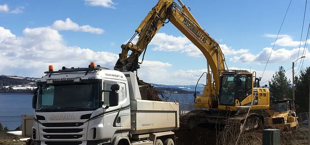 Gravemaskin fyller jord i lastebil fra Maskinkraft AS ved innsjø, med blå himmel og snødekte åser i bakgrunnen.