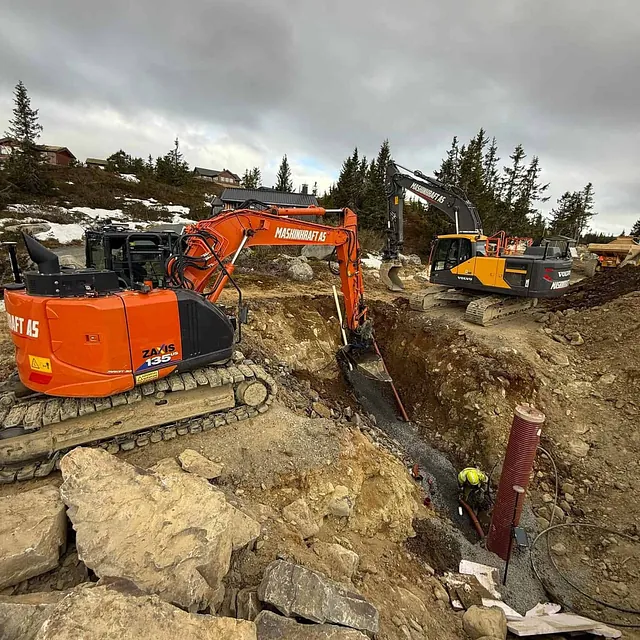 To gravemaskiner og en anleggsarbeider i grøft utfører rørlegging og graving i fjellterreng med hytter i bakgrunnen.