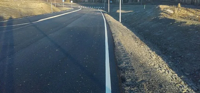 Nyasfaltert vei med hvit kantlinje og gangfeltmarkeringer, omgitt av grus og åpent landskap.