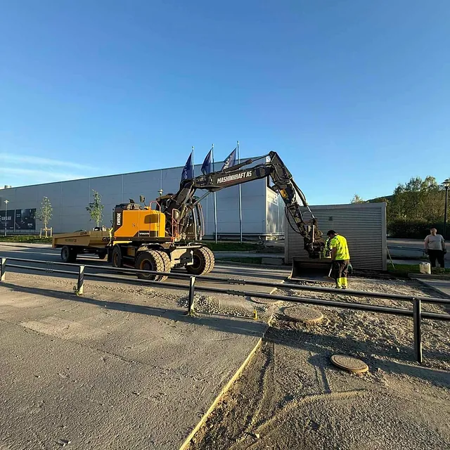 Gravemaskin på oppdrag ved fortau utenfor næringsbygg, med anleggsarbeider ved skuffen og kveldslys i bakgrunnen.