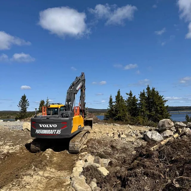 Volvo gravemaskin fra Maskinkraft AS arbeider på et steinlagt område med utsikt over innsjø og fjell.