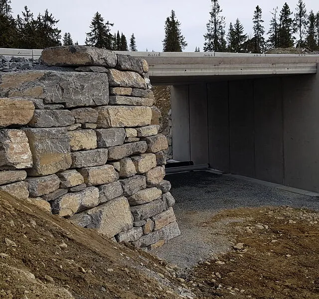 Undergang med støttemurer av naturstein under vei i skogsområde, med gruslagt gangsti gjennom.